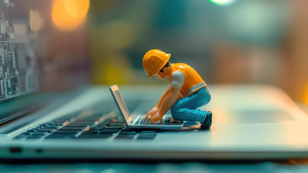 Miniature figure in a hard hat working on a small laptop resting on a real laptop keyboard